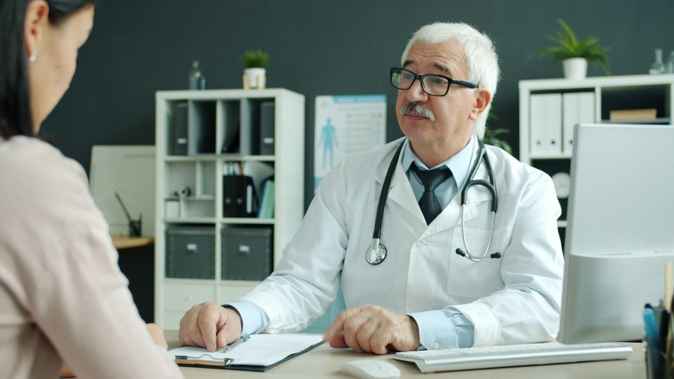 Médico de jaleco branco e estetoscópio atendendo uma paciente em um consultório moderno