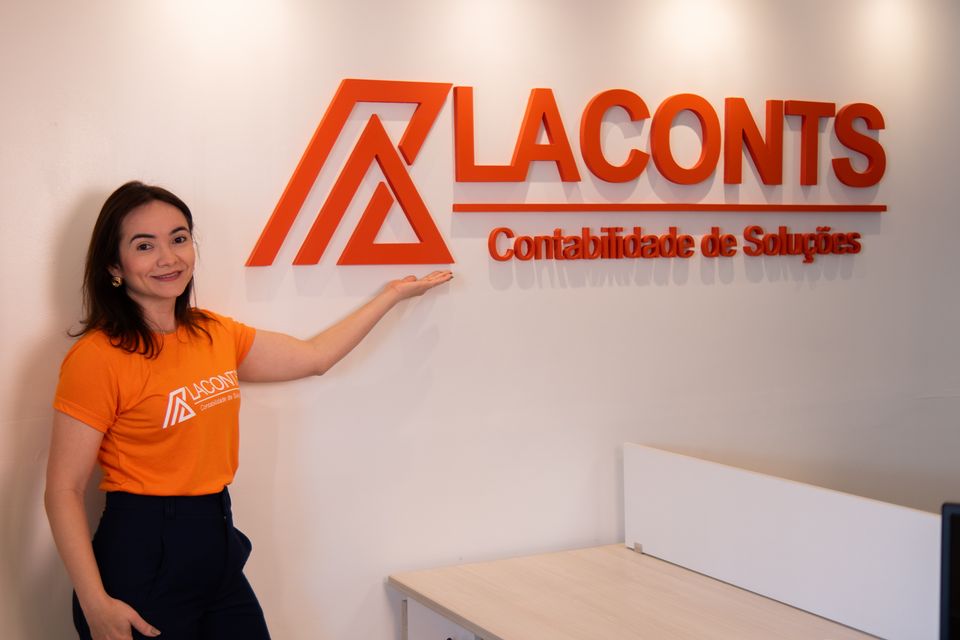 Pessoa da equipe Laconts, de camisa laranja, ao lado do logo da empresa na parede do escritório