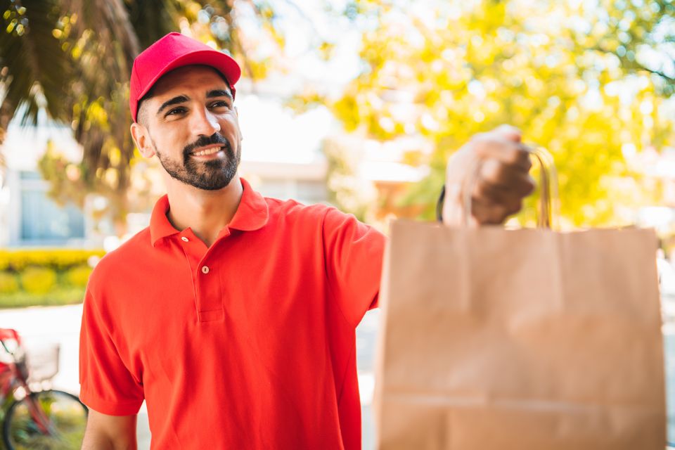 Entregador sorridente de camisa vermelha e boné, segurando uma sacola de entrega