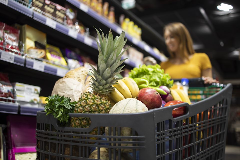 Carrinho de supermercado cheio de frutas e verduras, cliente escolhendo produtos ao fundo